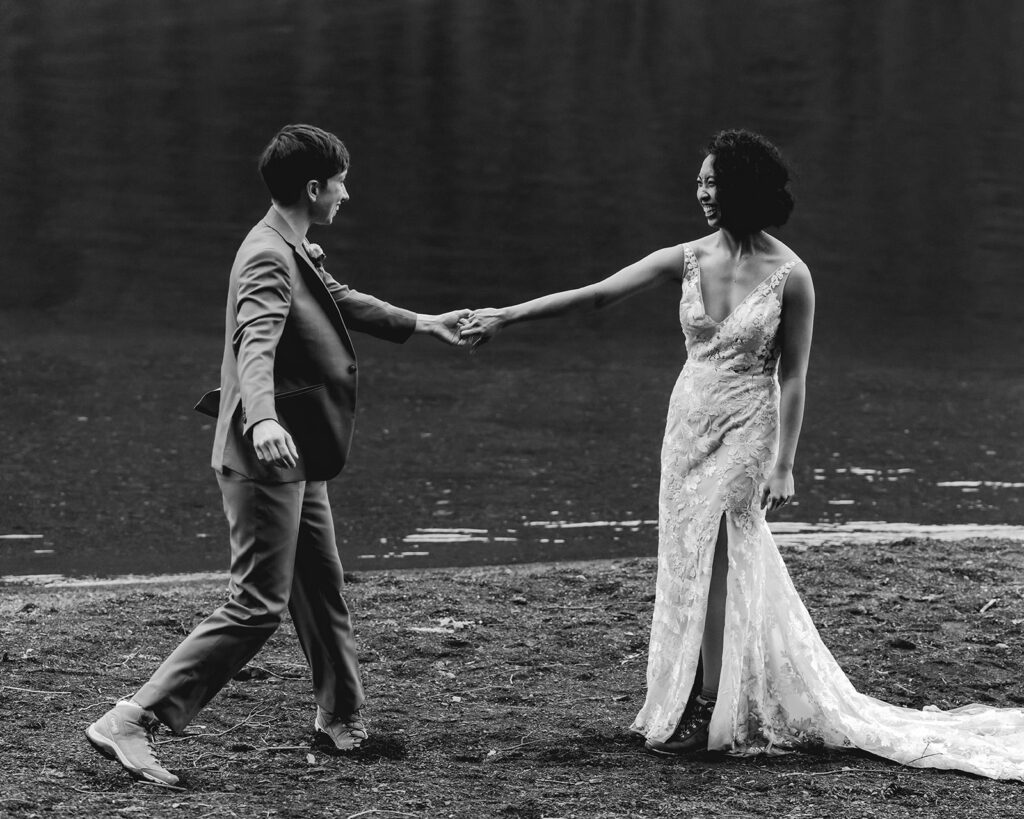 a black and white image of a couple in their wedding attire dance on the shore of the lake, smiling widely at each other during their lake crescent elopement. 