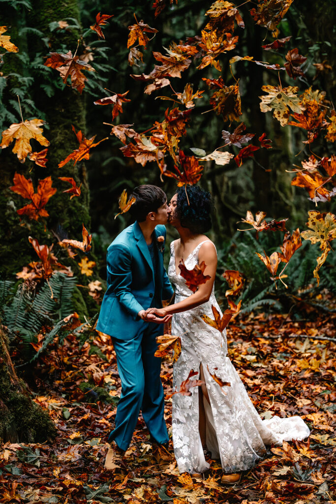 A couple in wedding attire kiss in the forest, bright colored autumn leaves fall all around them, almost in the shape of a heart during their Lake Crescent elopement