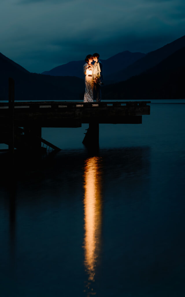 A couple in their wedding attire stand at the end of a dock, blue mountains in the background, the foreground is a reflection of the lantern light from the fixture they hold. it is just after sunset and the gold light illuminates them