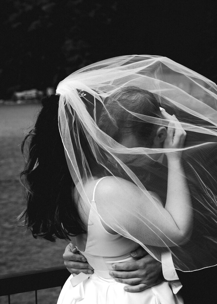 a bride and groom embrace in the wind during their lake crescent wedding. her veil billows over them as her groom kisses her shoulder