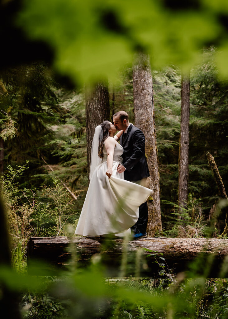 a couple in their wedding attire embrace in a lush forest. Framed by verdant leaves, they kiss and embrace during their lake crescent wedding.