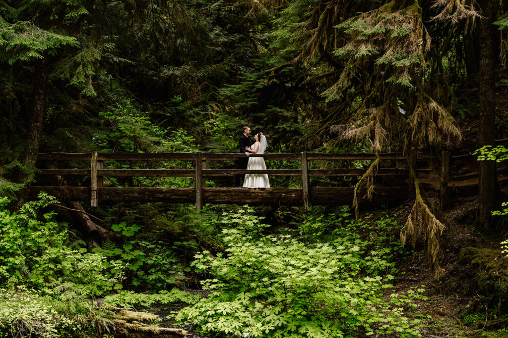 a bride and groom stand on a logged, wooden bridge. They are framed and surrounded by every shade of green the forest has to offer for their lake crescent elopement. 