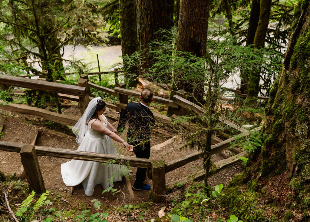 a bride and groom in their wedding attire walk down wooden steps on a forested trail. Green leaves surround them as they make their way to their lake crescent wedding location. 