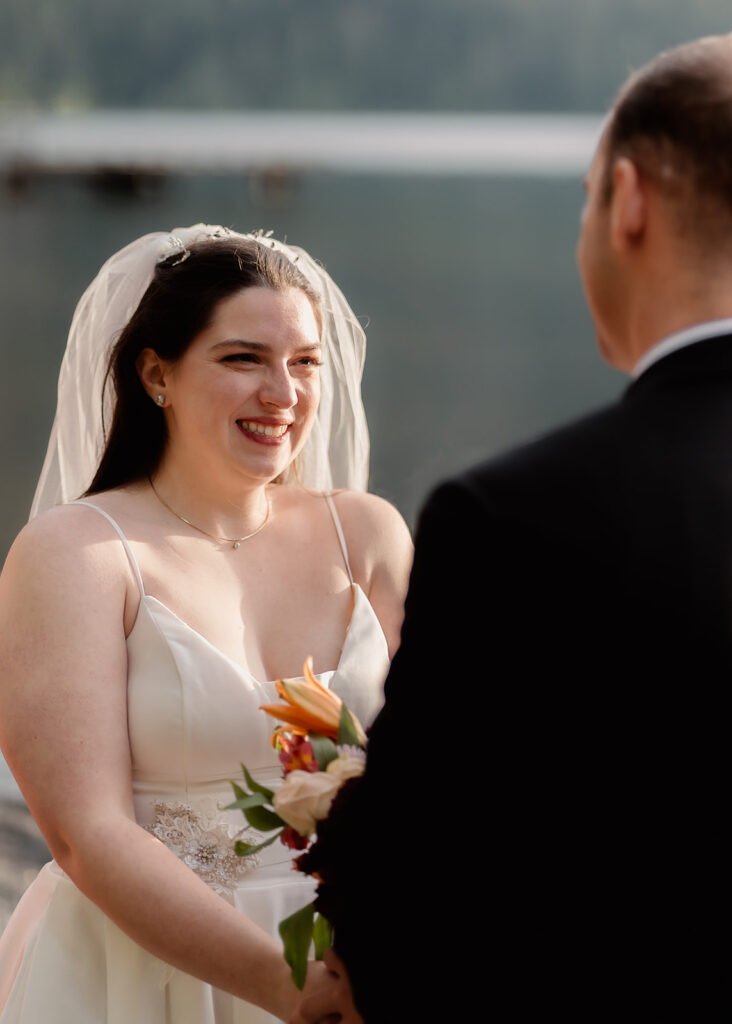 a bride smiles widely at her groom during their lake crescent wedding. she is glowing from joy and golden sunlight as this image is captured over her grooms shoulder