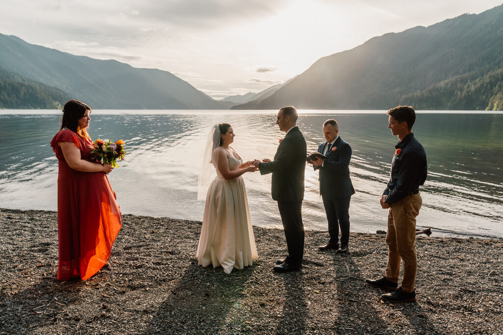 A lake crescent wedding ceremony painted with lens flare and dripping in golden, pink hues from the sun setting behind the lake. A bride and groom exchange rings as they are surrounded by their guests.