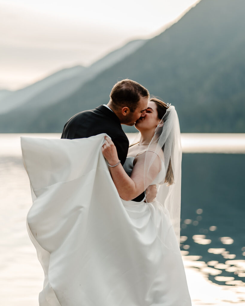 a bride and groom kiss in front of the sparkling, blue waters of lake crescent. We see the edges of dirst on her dress from their lake crescent wedding adventures.   