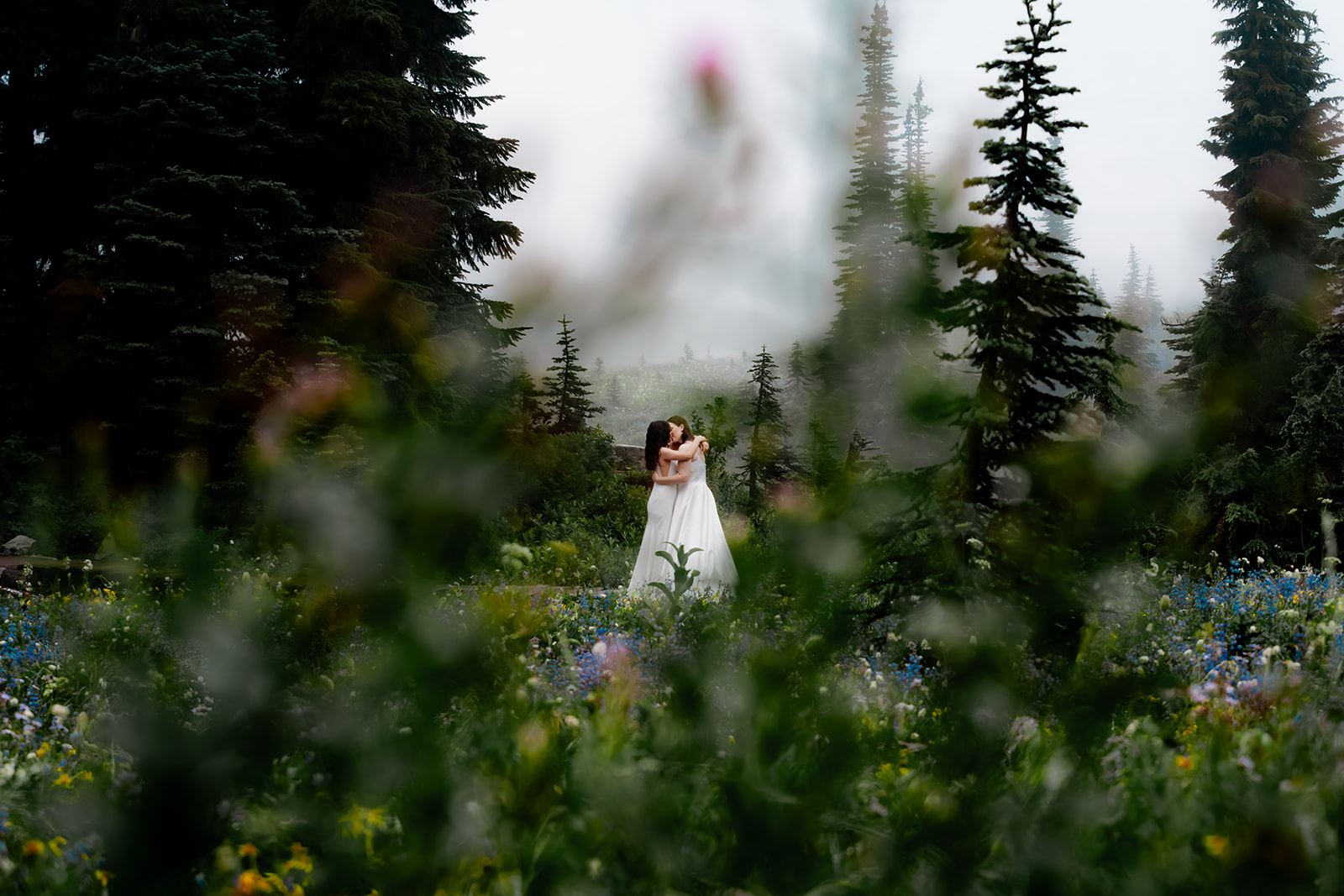Two brides embrace during their last minute elopement. the sky is gray and moody, but they are framed by colorful wildflowers and greenery