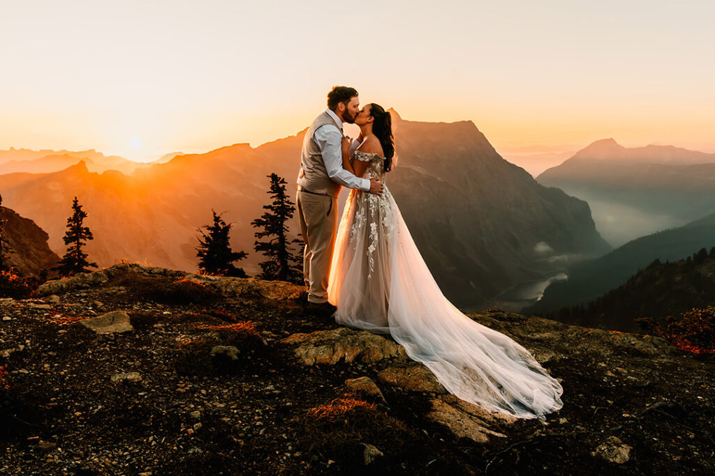 a couple in their wedding attire embrace and kiss at the top of a mountain during their north-cascades-elopement. it is clearly sunset and the landscape is painted with golden, pinky tones.